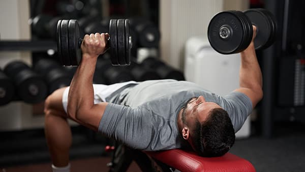 Lying on a weight bench, a man performs the dumbbell bench press exercise, with a dumbbell in each hand.