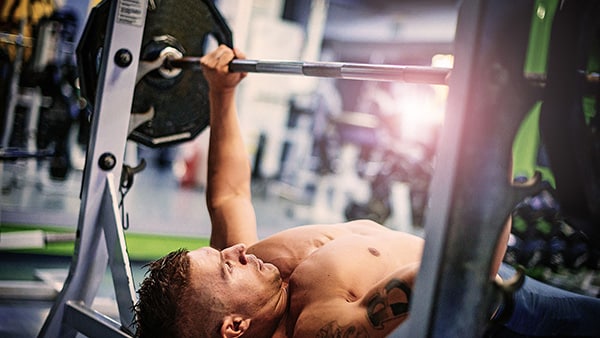 A shirtless man performs a bench press exercise in a gym.