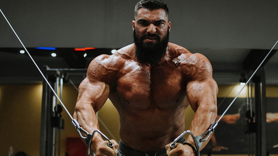 A shirtless man performs a pectoral isolation exercise with a pulley in a weight room.