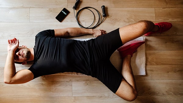 Un sportif est allongé sur une serviette posée sur le sol, avec un smartphone et une corde à sauter à côté de lui, visiblement fatigué par sa séance d'entraînemen.