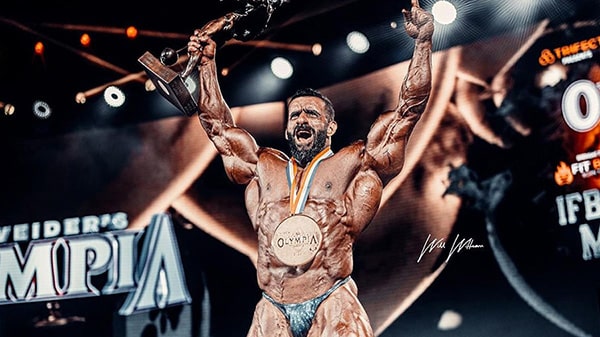 Hadi Choopan celebrates his victory at Mr. Olympia 2022, arms in the air, the medal around his neck and the trophy in his right hand.