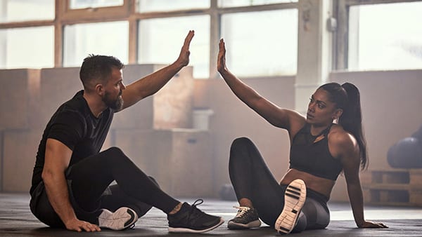 In a fitness room, two sportsmen (a man and a woman) are sitting on the floor and slapping their hands.