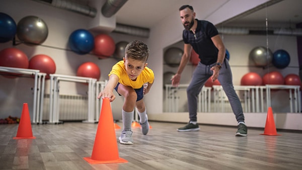 A child performs fitness exercises in a gym, supervised by a sports coach.