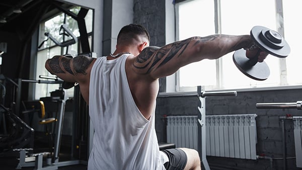 Un homme de dos, assis sur un banc de musculation, fait des élévations latérales aux haltères pour muscler ses épaules.