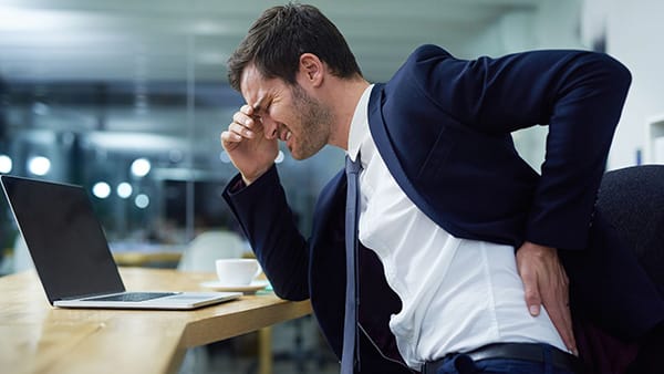 A man in a suit and tie, sitting at a desk in front of a computer, suffers from lower back pain and touches his lumbar region with his left hand.