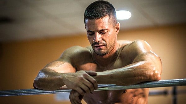 A bodybuilder, shirtless, is leaning with his elbows on a bar during his recovery time.