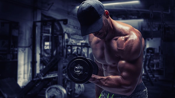 Un homme torse nu, avec une casquette sur la tête, réalise un exercice de musculation pour les biceps avec des haltères dans une salle de fitness.