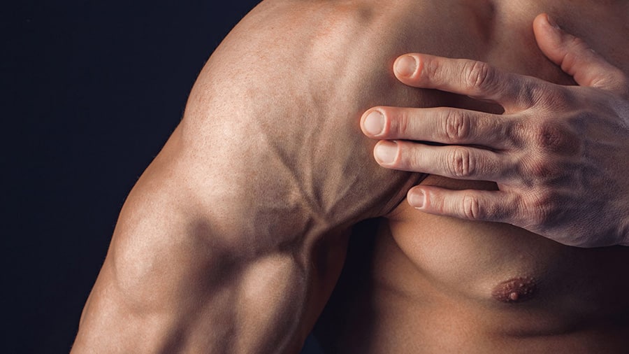 A shirtless man, victim of aches and pains, holds his right pectoral muscle with his left hand.