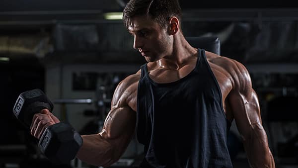 A man in a tank top performs the biceps curl exercise in a weight room.