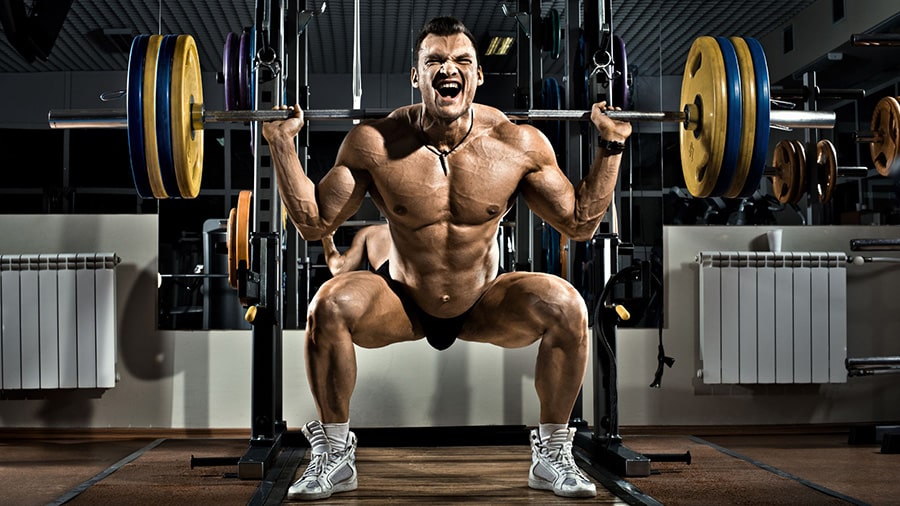 A bare-chested weightlifter performs the free bar squat using the pyramid method in a gym.