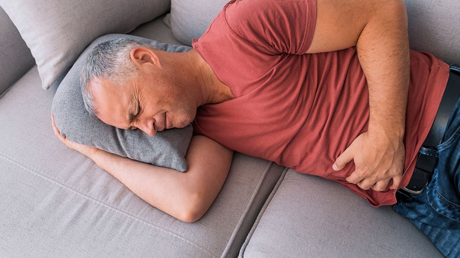 A man, dressed in jeans and a red t-shirt, is lying on a couch and suffers from stomach pains because of his perineum.