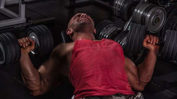 A sportsman in a red tank top does a dumbbell bench press on a weight bench to build up his pectoral muscles.