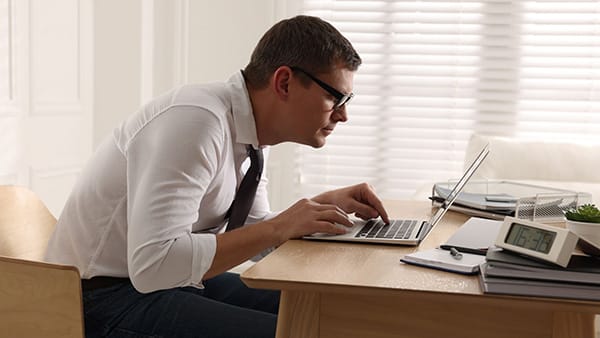 A man in a shirt and tie is working on a computer, sitting at a desk with a bad posture (sunken back).