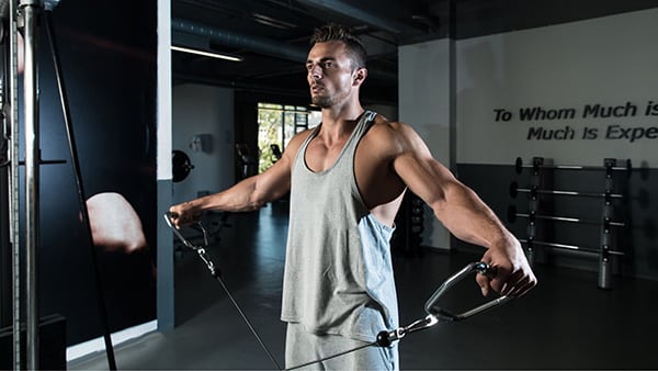 A bodybuilder wearing a grey tank top performs low pulley lateral shoulder raises in a gym.