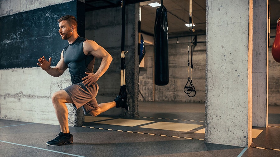 A man in shorts and a tank top performs a TRX lunge exercise in a gym to strengthen his stabilizing muscles.