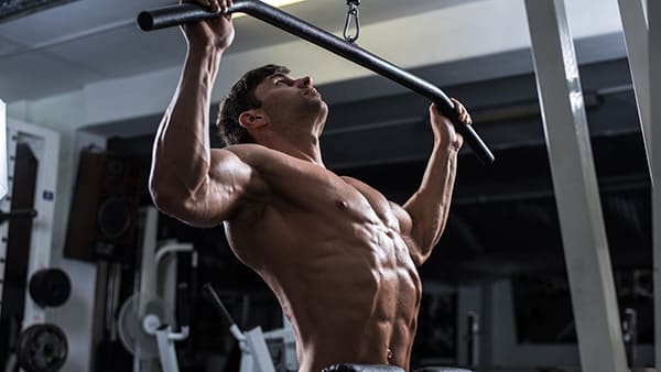 A shirtless man in a gym performs the vertical pull-up exercise to build back muscles with a wide grip.