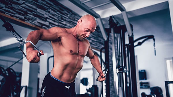 A shirtless man in a fitness center performs a pectoral muscle-building exercise using a pulley.