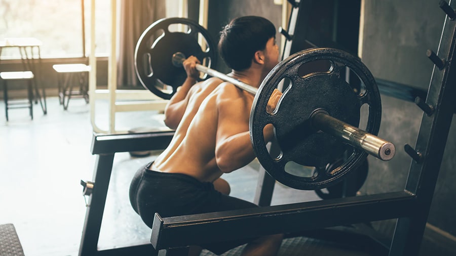 A shirtless man in a gym applies the stato-dynamic method to the bar squat exercise.