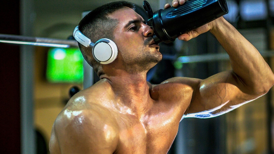 A shirtless man, sitting on a weight bench in a gym with headphones on, drinks water mixed with dextrose in a shaker.