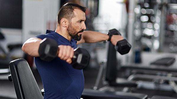 A man in a weight room does a lateral raising exercise with dumbbells in each hand.