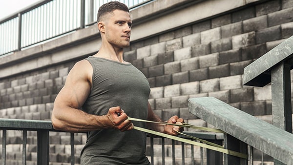 A man in a tank top performs a pulling exercise with a fitness elastic as part of a bodybuilding warm-up.