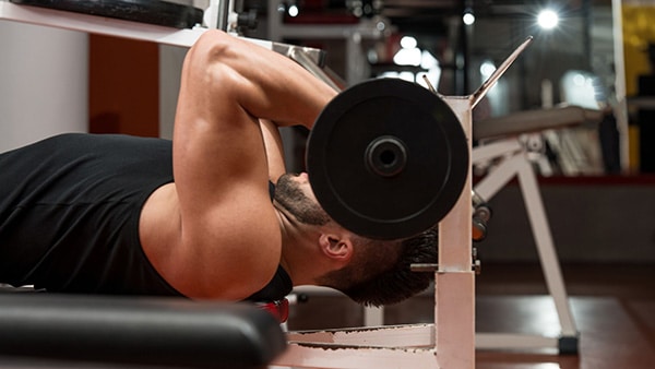 A bodybuilder in a tank top performs the barbell-to-front triceps exercise in a gym.