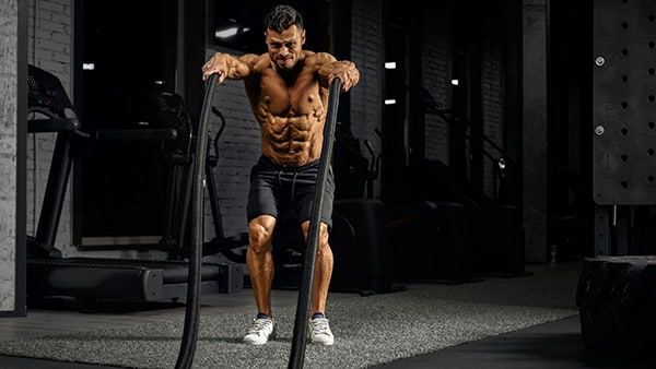 A shirtless man performs a muscle-building exercise with a battle rope in a fitness center.