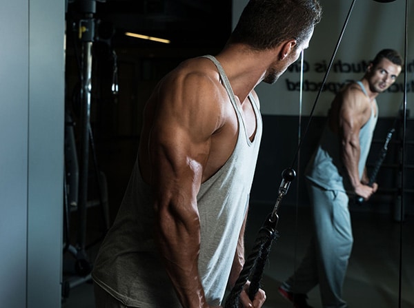 Un pratiquant de musculation en débardeur effectue des extensions à la poulie haute dans une salle de sport.