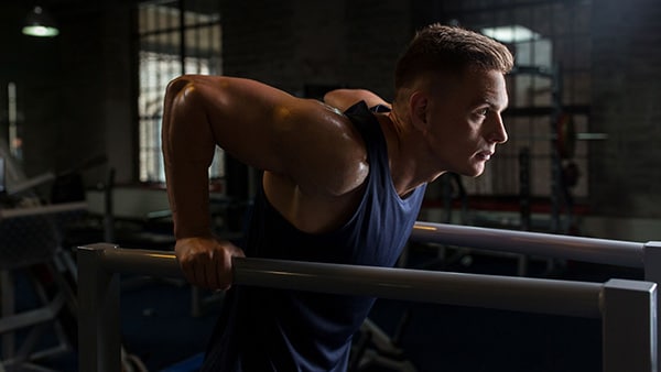 A bodybuilder in a tank top performs dips on parallel bars.