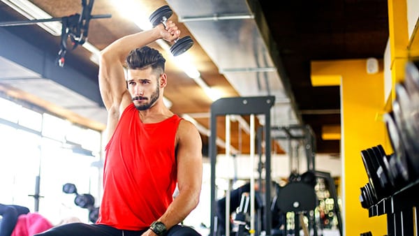 A bodybuilder in a red tank top stretches the back of his neck with a dumbbell in his left hand.