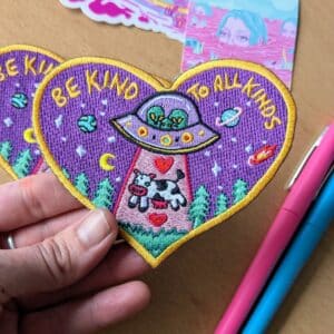 Heart-shaped embroidered patch with a psychedelic design featuring an alien spaceship beaming up a cow, surrounded by planets, stars, and trees on a purple background. The patch includes the text 'BE KIND TO ALL KINDS' in yellow letters, bordered by a golden edge. Perfect for jackets, backpacks, or hats.