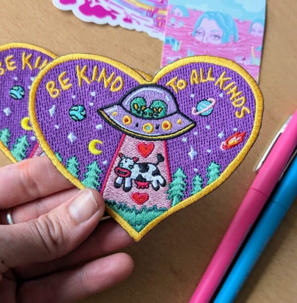 Heart-shaped embroidered patch with a psychedelic design featuring an alien spaceship beaming up a cow, surrounded by planets, stars, and trees on a purple background. The patch includes the text 'BE KIND TO ALL KINDS' in yellow letters, bordered by a golden edge. Perfect for jackets, backpacks, or hats.