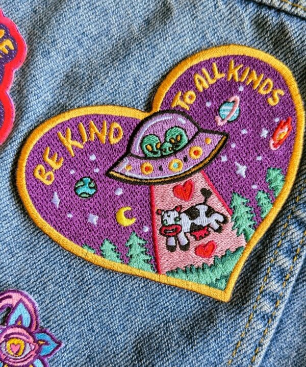 Heart-shaped embroidered patch with a vibrant design featuring an alien spaceship beaming up a cow, surrounded by planets, stars, and trees on a purple background. The patch includes the text 'BE KIND TO ALL KINDS' in yellow letters, refering to peace and to go vegan. Perfect for jackets, backpacks, or hats.