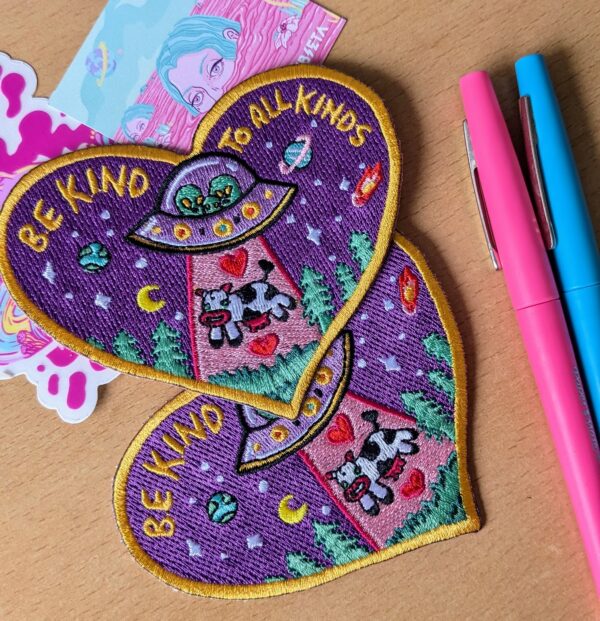 Heart-shaped high quality embroidered patch with a pop design featuring 3 cute aliens in an ufo beaming up a cow, surrounded by planets, stars, and trees on a purple background. The patch includes the text 'BE KIND TO ALL KINDS' in yellow letters, bordered by a golden edge. Perfect for jackets, backpacks, or hats.