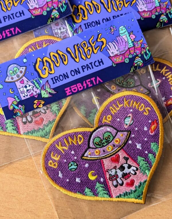 Quirky heart-shaped embroidered patch with a fun and eclectic design featuring an alien spaceship abducting a cow, set against a purple background with cosmic elements like planets and stars. The patch reads 'BE KIND TO ALL KINDS' in bold yellow lettering and is outlined in a stylish golden border. Ideal for adding a touch of unique Portland flair to jackets, backpacks, or hats