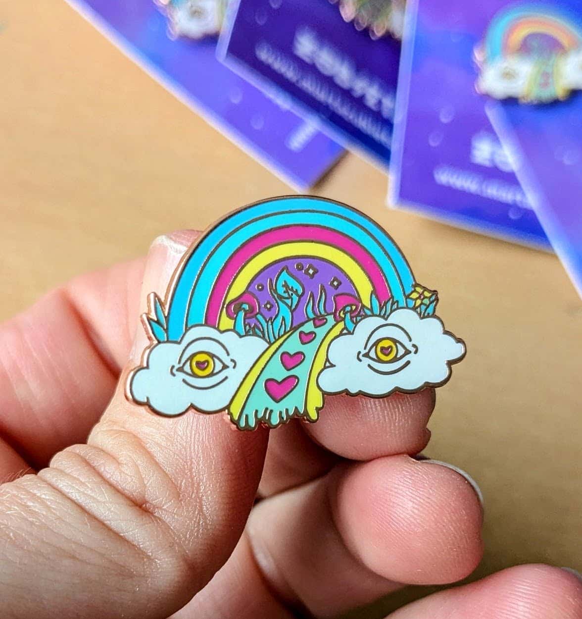 Colorful enamel pin featuring a quirky rainbow emerging from two cartoon clouds with eyes, leading to a path decorated with pink hearts and surreal elements. Perfect for adding a touch of quirky, alternative flair to jackets, bags, or hats, and embracing the unique and artistic spirit of the queer community