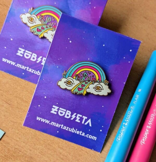 Colorful enamel pin featuring a vibrant rainbow emerging from two whimsical clouds with eyes, leading to a path decorated with pink hearts and surreal elements. Perfect for adding a touch of quirky, alternative flair to jackets, bags, or hats, and embracing the unique and artistic spirit of Bristol