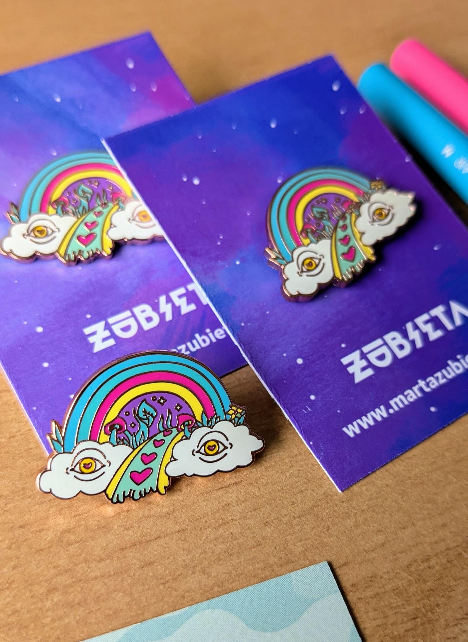 Colorful enamel pin featuring a vibrant rainbow emerging from two whimsical clouds with eyes, leading to a path decorated with pink hearts and surreal elements. Perfect for adding a touch of quirky, alternative flair to jackets, bags, or hats, and embracing the unique and artistic spirit of the lgbt+ community