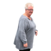 En kvinde med kort gråt hår og briller, iklædt en lun cardigan fra OPSKRIFT - MODEL ASTRID - CARDIGAN MED SNONINGER kollektionen, står på en hvid baggrund. Hendes øjne funkler af stille tilfredshed, mens hun smiler varmt, efter at have mestret sin strikkemaskine.