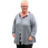 En smilende ældre kvinde med kort hvidt hår bærer briller og OPSKRIFT - MODEL ASTRID - CARDIGAN MED SNONINGER, en grå cardigan med knapper, over en sort top med blomstermønstre. Den hvide baggrund fremhæver subtilt de indviklede designs, der minder om strikkemaskines håndværk.