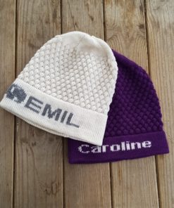 To strikbeanies hviler på en træoverflade. På den fløde hue på toppen står der "EMIL" med grå bogstaver, mens den lilla under den siger "Caroline" i hvidt. Begge er lavet af bæredygtigt garn, der viser et tekstureret, honeycomb-mønster lavet med præcision på en strikkemaskine.