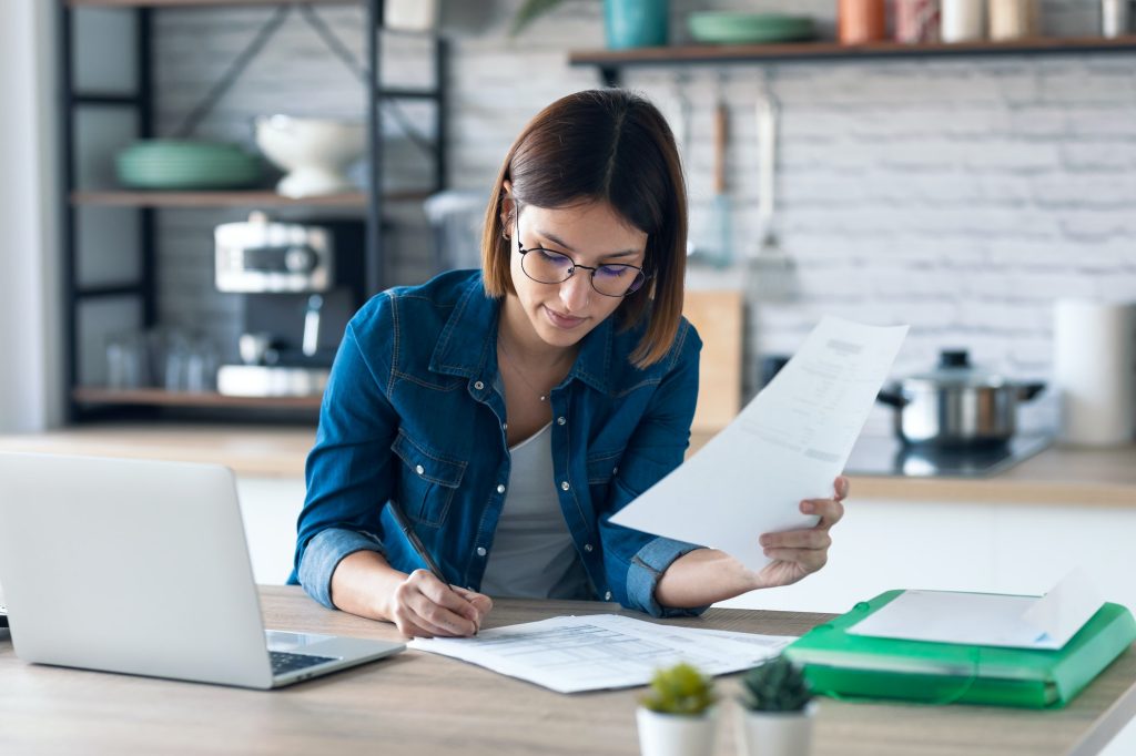 young woman working with computer while consulting some invoices and documents in the kitchen