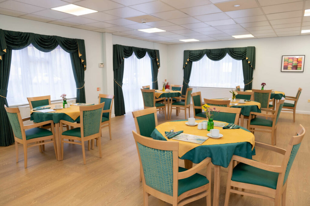 Dining area with laid wooden tables and chairs. The table clothes and chair backs and seats are blue/green colour.