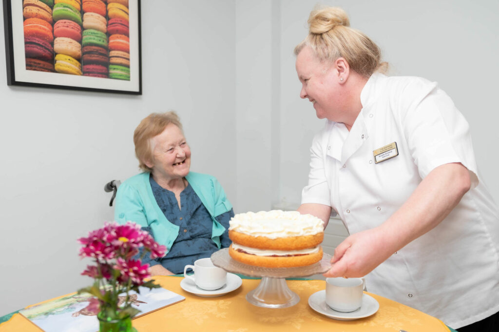 Member of staff smiling with an elderly resident across the table which has a cake on it.