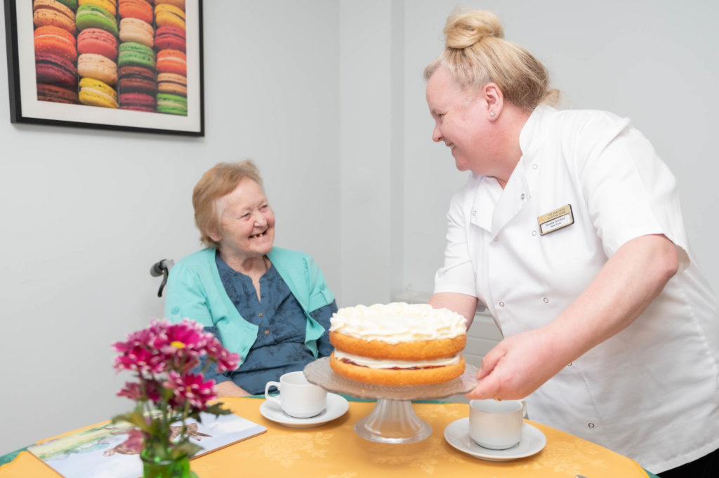 A member of staff smiling with an elderly resident with a cake in the middle of the table.