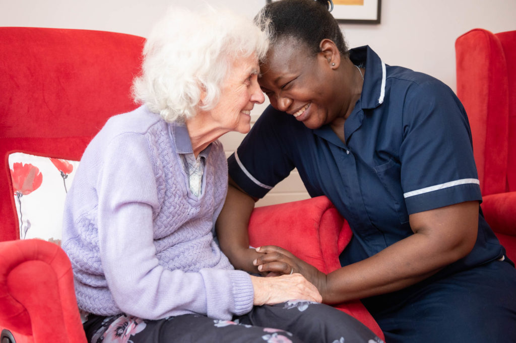 A member of staff and resident holding hands and pressing their heads together whilst smiling.