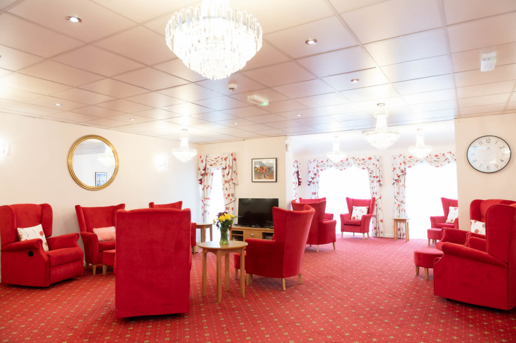 The lounging area which has red arm chairs, a red patterned carpet, white walls and red and white curtains.
