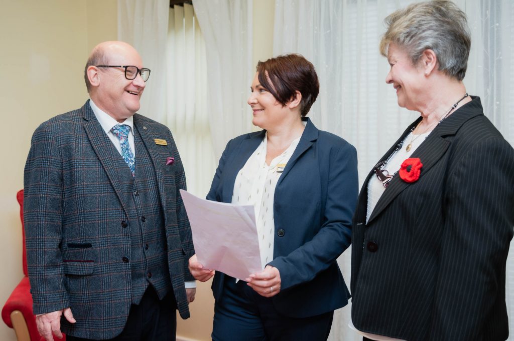 Member of staff holding a piece of paper stood between two smiling individuals.
