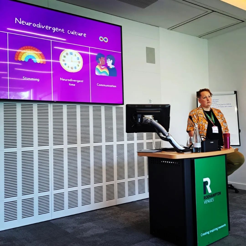 A person in an orange shirt sits on the far side of a lectern. A screen behind them shows a magenta image with brightly coloured icons