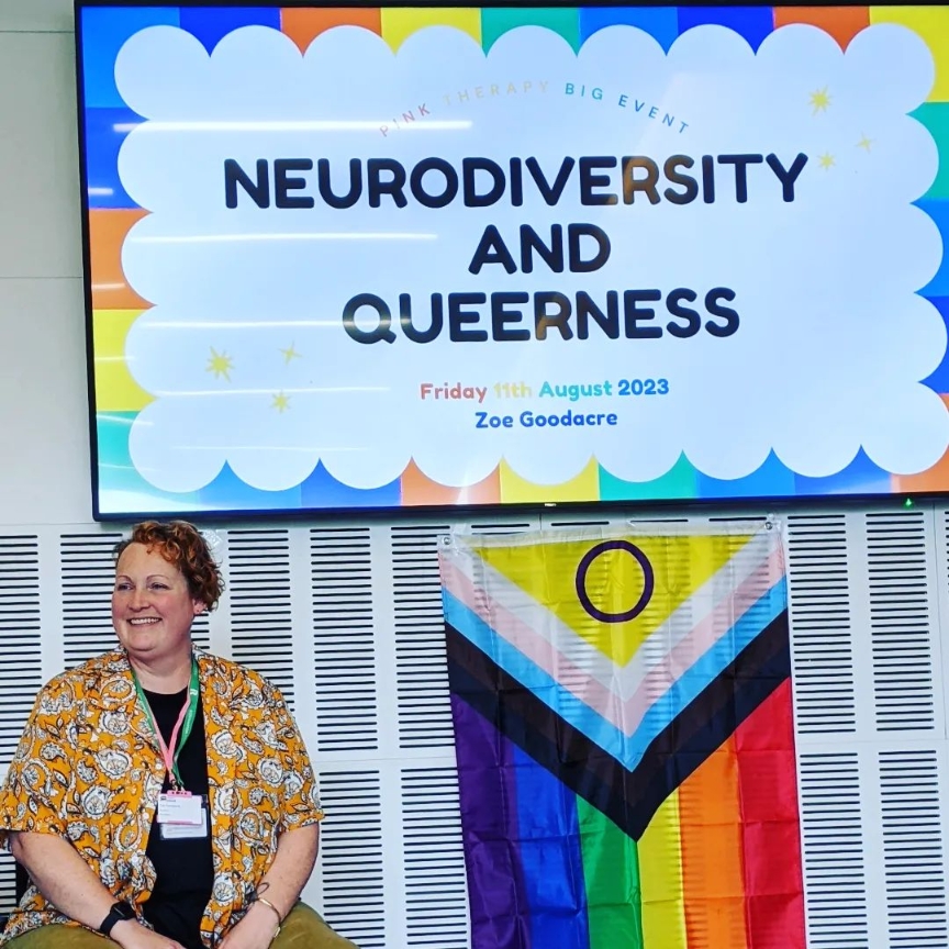A person in an orange paisley shirt sits smiling in front of a progress pride flag. A screen in the background reads "Neurodiversity and Queerness"